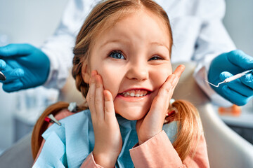 Humorous portrait of a cute little girl with pigtails sitting in a dental chair, looking at the camera and grimacing. Behind, a doctor in gloves holds examination tools. - Powered by Adobe