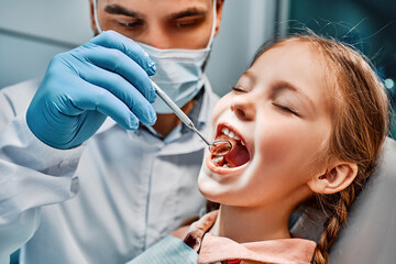 Checking teeth at a children's dentist. Close-up image of a doctor examining her teeth with a mirror, a small female patient sitting in a chair with her eyes closed.