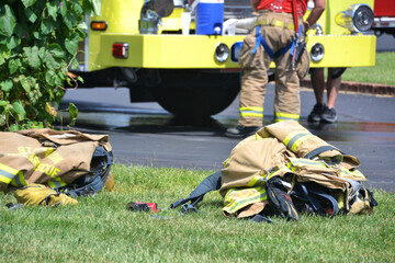 Protective firefighter gear lying on the ground with firemen and a firetruck in the background.
