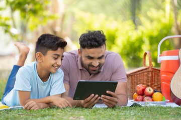 Happy smiling indian father with son using digital tablet during weekend picnic at park - concept...