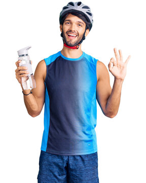 Handsome hispanic man wearing bike helmet and holding water bottle doing ok sign with fingers, smiling friendly gesturing excellent symbol