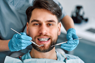 Cropped portrait of handsome man with braces sitting in dental chair looking at camera and smiling. Behind, a doctor in gloves holds examination tools.