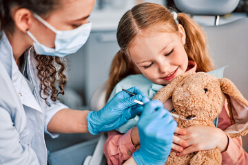 At the doctor's appointment. A child is sitting in a dental chair and holding a toy rabbit and a nurse is playfully examining the toy. Approach to the patient.