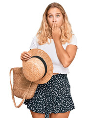 Young blonde girl holding summer hat and wicker handbag covering mouth with hand, shocked and...