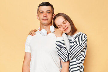 Smiling calm woman embracing serious man wearing casual outfits standing isolated over beige background female closed eyes felling love, guy looking at camera.