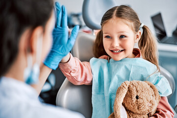 A candid emotional photo of a child sitting in a dental chair, holding a toy rabbit and cheerfully giving a high-five to the nurse. Children's dentistry.