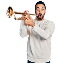 Young hispanic man with beard playing trumpet afraid and shocked with surprise and amazed expression, fear and excited face.