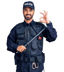 Young hispanic man wearing police uniform holding baton doing ok sign with fingers, smiling friendly gesturing excellent symbol