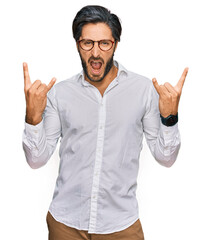 Young hispanic man wearing business shirt and glasses shouting with crazy expression doing rock symbol with hands up. music star. heavy concept.