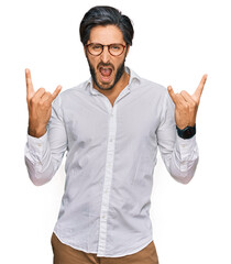 Fototapeta premium Young hispanic man wearing business shirt and glasses shouting with crazy expression doing rock symbol with hands up. music star. heavy concept.