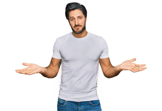 Young hispanic man wearing casual white t shirt clueless and confused expression with arms and hands raised. doubt concept.