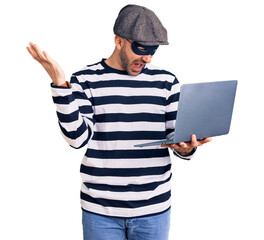 Young handsome man wearing burglar mask using laptop celebrating victory with happy smile and winner expression with raised hands