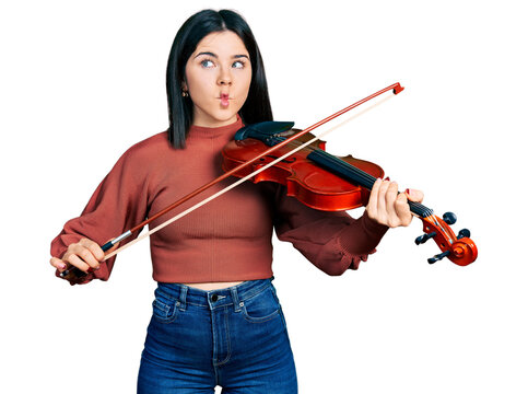 Young brunette woman with blue eyes playing violin making fish face with mouth and squinting eyes, crazy and comical.