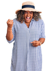 Young african american plus size woman wearing summer hat angry and mad raising fist frustrated and furious while shouting with anger. rage and aggressive concept.
