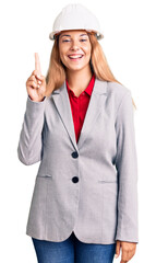 Beautiful young woman wearing architect hardhat showing and pointing up with finger number one while smiling confident and happy.