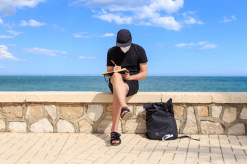 Male writer in mask writing in notebook on embankment
