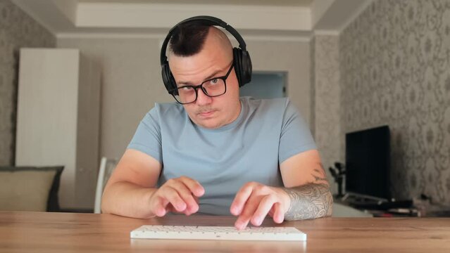 Caucasian male nerd in glasses with tattooed hands sitting at desk and working on computer. Geek funny man programmer looking at camera and typing on modern keyboard, listening music in his headphones