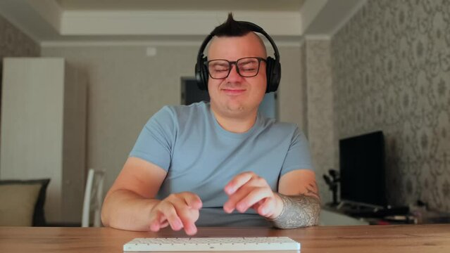 Geek funny man programmer fast typing on modern keyboard, shaking his head listening music in his headphones. Caucasian male nerd in glasses with tattooed hands sitting at desk and working on computer