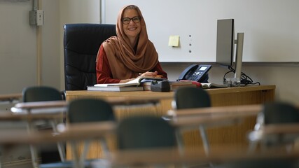 Portrait of happy smiling woman teacher wearing headscarf in school classroom desk looks up and gives thumbs up.