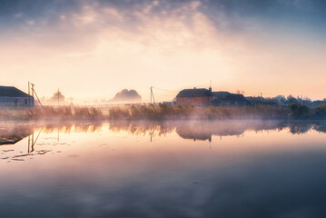 Beautiful morning scene in tranquil village with a lake in the mist at sunrise. Countryside with houses in the sun rays at dawn.