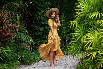 Female tourist girl with fashionable hat poses elegantly in lush green jungle. Young woman dancing...