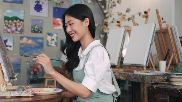 Woman paints picture on canvas with oil paints in her studio. Female artist painting picture in workshop. Young female artist painting on a canvas with a paintbrush and holding a color palette at home