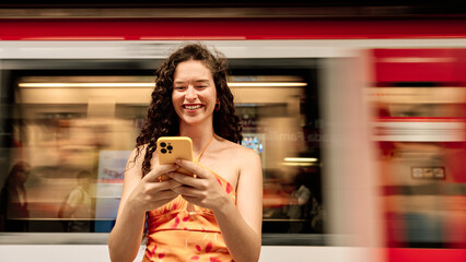 young woman using smartphone while subway passes behind her - transportation and commute concept - obrazy, fototapety, plakaty