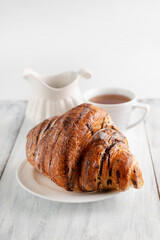 On a gray table is a blurry image of a croissant, a cup of tea and a jug of milk.