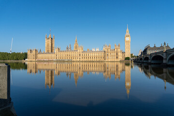 Photo of the entire English Parliament next to Big Ben. Photo taken from the other side of the river, with the reflection of the entire parliament in the river water. On a sunny day.