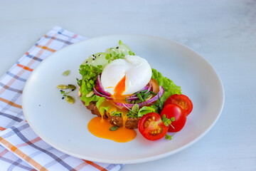 Runny poached egg on wholemeal bread with salad, tomato, red onion, lettuce and cucumber. Decorated with cherry tomatoes, microgreens, sesame and pumpkin seeds on white plate on cotton chessboard
