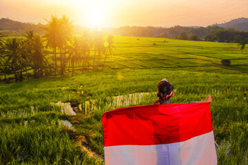 boy waving Indonesian red and white flag with a view of rice fields in the afternoon, against the...