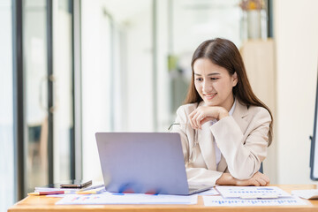Asian female entrepreneur working in finance at home office analyzing financial graphs on documents with laptop on table in office