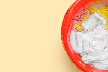 Plastic basin with dirty clothes, water and foam on yellow background