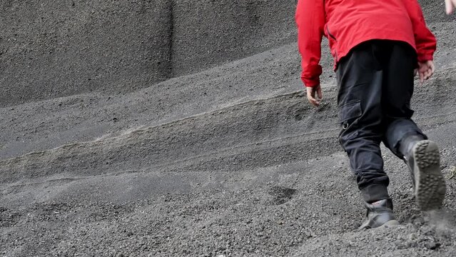 A young boy and girl in red jackets and a backpack climb uphill to a gray loose rock made of volcanic ash and slag of an ancient extinct volcano. Teen activity