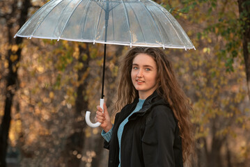 Portrait of smiling beautiful woman with long hair under transparent umbrella. Cute girl stands on alley of fall park