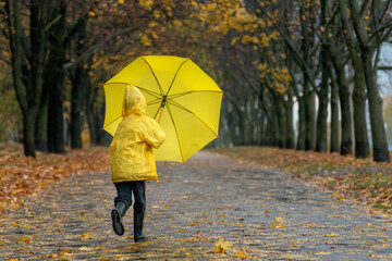 Child in yellow raincoat with an umbrella in his hands runs through rainy autumn park. Rear view