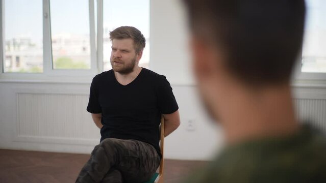 Panning shot of stressed bearded military soldier in camouflage uniform having conversation during post war group support session therapy. Concept of mental health, psychotherapy, social issues.