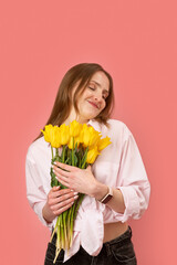 Happy young european lady holds bouquet of yellow tulips with closed eyes, pink background. Gift to women day. Mother day. March 8.