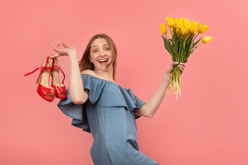 Happy fun young woman in summer sundress stands barefoot holds sandals and bouquet of flowers....