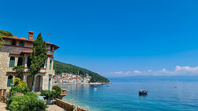A panoramic view of the shore along Moscenicka Draga, Croatia. There is a small town located on the shore of the Mediterranean Sea. Few boats crossing the calm sea. Green hills in the back. Sunny day.
