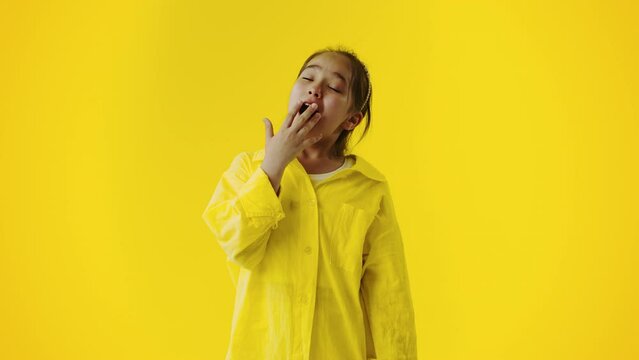 Tired and bored young girl yawns widely, absently covering her mouth with a hand on a yellow background. Concept of the lack of interesting activities for the child