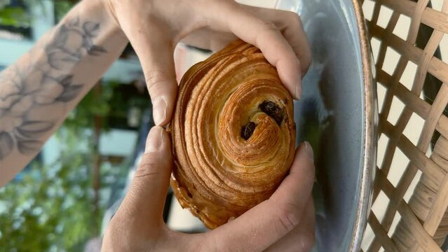 Close-up of female hands tearing fresh Pain au chocolat pastry into halves on the cafe terrace. City atmosphere of a warm summer day in bokeh background. Lifestyle vertical video.