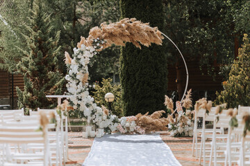 The wedding arch in the park is made of fresh flowers and dry reeds. Away wedding ceremony. White chairs for the ceremony