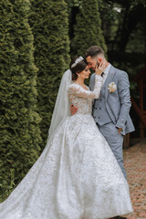 Naklejka premium European wedding couple in the park. The bride in a beautiful dress with sleeves and a crown on her head. Groom with a black beard of Caucasian appearance in a classic suit