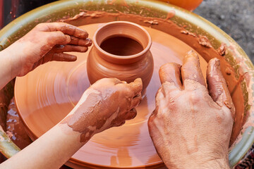 Teaching a child to make pottery from red clay. Clay pottery hands close up working on wheel handcrafts. Clay crockery making. Working with red clay. Hands and fingers of potter and child in clay.