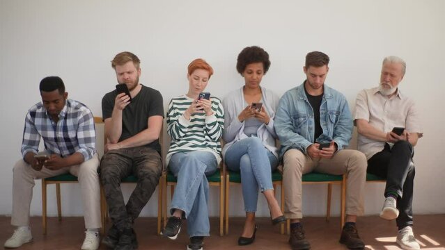 Wide shot of multicultural and different ages people sitting on chairs in row waiting for job interview using smartphones. Handsome young man standing up and walking away. Shooting in slow motion.