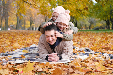 Family day in autumn park. Father with daughters lies on a blanket and plays. Daughters tickle father
