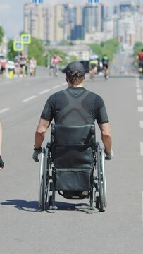 Vertical Screen: disabled athlete on wheelchairs taking part in street race together with runners, modern buildings in the distance