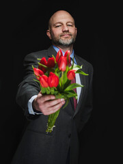 Man holding fresh red tulip bouquet in his hands. Model is bald with grey beard, wearing classic grey suit. Handsome male with special gift. Making up concept. Dark color background.