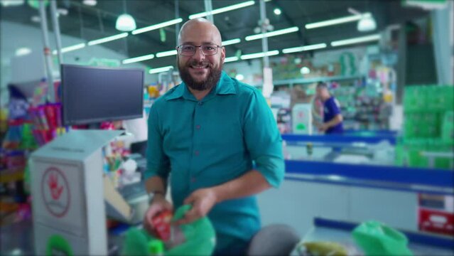 One happy Brazilian employee of supermarket at checkout scanning products and putting into plastic bags while smiling at camera. Grocery Market scene in South America
